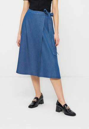 Nő, kék farmer megkötős szoknyát visel, amely a derekán meg van kötve, fekete felsővel és fekete, arany lánccal díszített, tömör sarkú, lakkozott bőr mokaszinokkal.