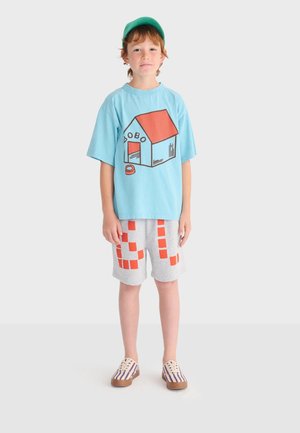 Boy wearing a green cap, blue t-shirt with a doghouse graphic, gray shorts with orange squares, and striped sneakers standing on a white background.