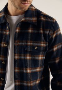 Laivastonsininen flanellipaita, jossa ruskea ja valkoinen ruutukuvio. Siinä on kaulus, nappikiinnitys edessä ja rintatasku, jossa on nappisuljin.