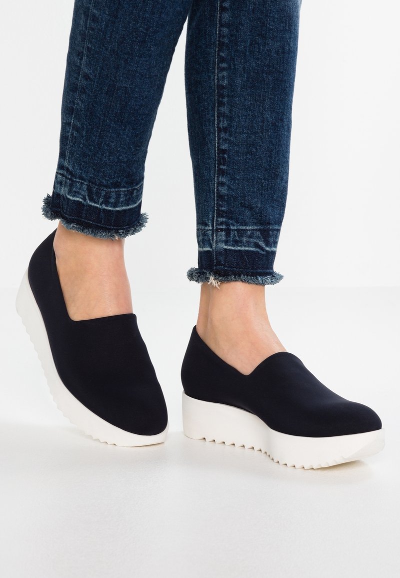 Vrouw in donkere instapschoenen met een platform en dikke witte zolen, gecombineerd met versleten donkerblauwe spijkerbroek.