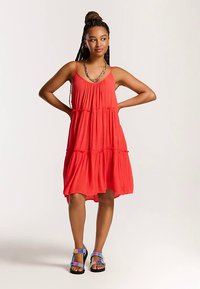 Red sleeveless dress with tiered design, made of lightweight fabric, features a flowy shape and detailed ruffle accents. Worn with multicolored sandals.