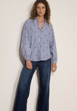 Frau trägt eine lockere blau-weiß gestreifte Bluse mit kleinen Herzmustern und weite dunkelblaue Jeans, steht vor einer schlichten Wand.
