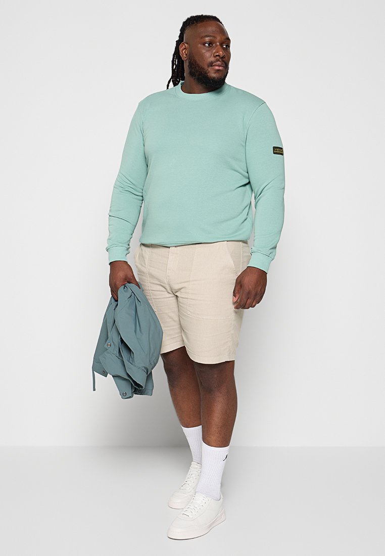 Miętowa, długą rękawą koszula z okrągłym dekoltem, jasnobeżowe szorty, białe sneakersy, trzymając jasnoniebieską kurtkę. Prosty design, miękka tekstura.