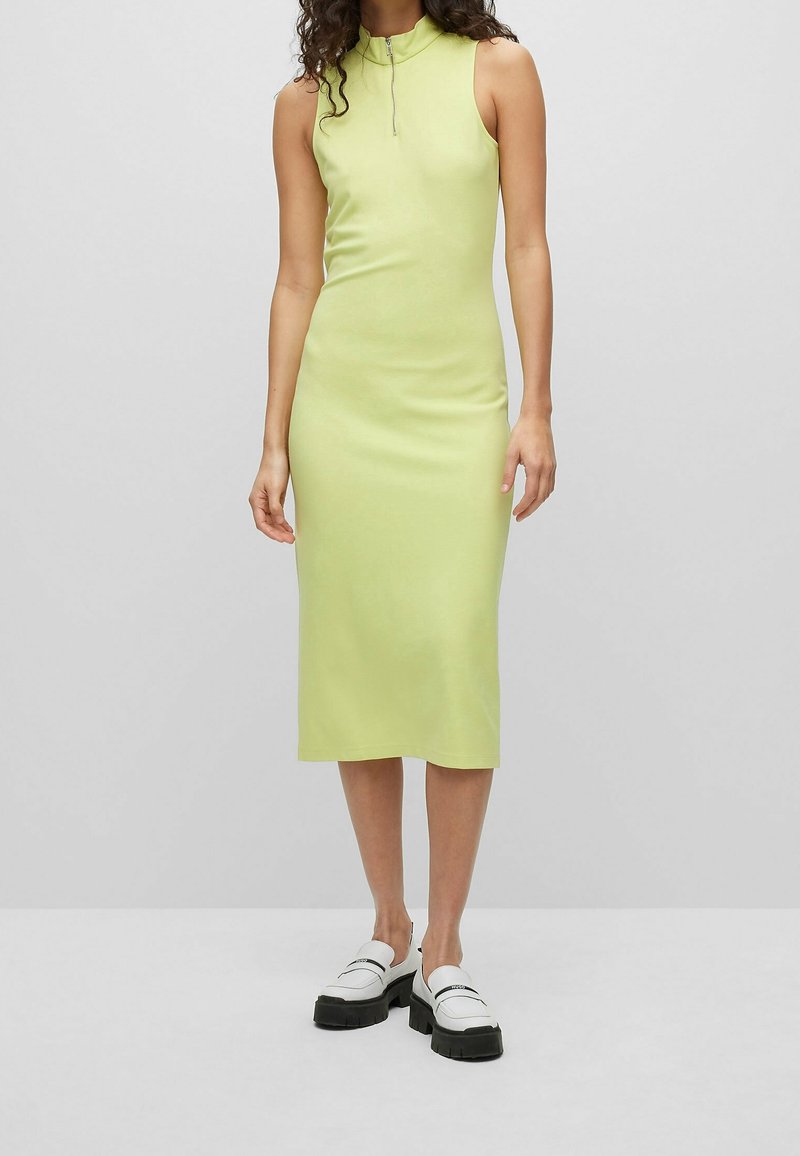 Knielanges, ärmelloses Kleid aus leicht limettengrünem Stoff mit hohem Kragen und einem Reißverschluss auf der Vorderseite. Kombiniert mit auffälligen weißen Slippern.