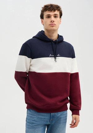 Giovane uomo con felpa con cappuccio a blocchi di colore blu navy, bianco e bordeaux con coulisse e jeans blu, in piedi davanti a uno sfondo semplice.