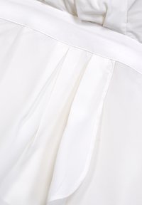 Vit blus med ett mjukt tyg, veck längs sömmen och en platt krage. Designen är enkel med rena linjer och minimala detaljer.