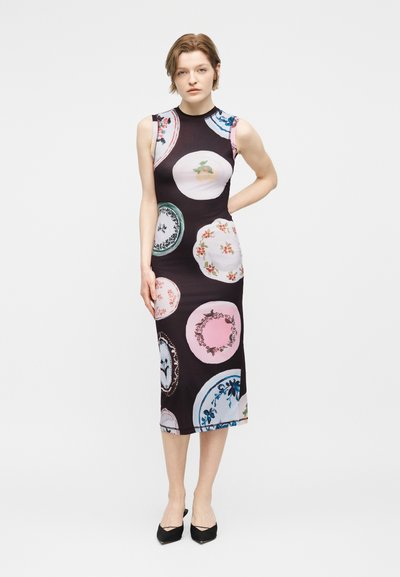 Γυναίκα που στέκεται φορώντας ένα αμάνικο, εφαρμοστό μαύρο μίντι φόρεμα με πολύχρωμα κυκλικά λουλουδένια και φρουτώδη σχέδια, συνδυασμένο με μαύρα μυτερά παπούτσια.