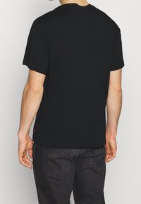 Mężczyzna noszący prostą czarną koszulkę z krótkim rękawem i ciemne dżinsy, pokazany od tyłu na jednolitym jasnym tle.