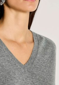 Frau in einem grauen V-Ausschnitt-Wollpullover und einem großen dekorativen Ohrring, von Kinn bis Schultern vor einem schlichten Hintergrund gezeigt.