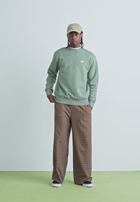 Groene sweatshirt met een klein wit logo, gecombineerd met bruine geruite wijde broek en zwarte sneakers. Model draagt een beige pet.