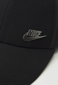 Sort kasket lavet af tekstureret stof med et sølvfarvet Nike-logo foran, har en buet skygge og synlige syninger.