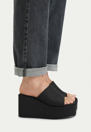 Piedi che indossano sandali platform neri aperti sulle dita, abbinati a jeans grigio scuro risvoltati, su uno sfondo chiaro uniforme.