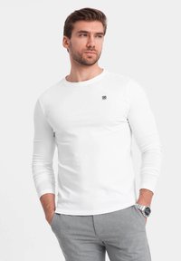 Langarmige witte T-shirt van zachte, rekbare stof met een ronde hals. Kleine logodetail op de linkerkant van de borst. Model draagt een grijze broek.