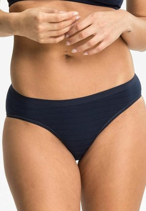 Woman in navy blue bikini briefs with hands resting on lower ribs and part of a black swimsuit top visible.