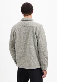 Grijze textuur wol shirtjack met een neklap, knoopsluiting aan de mouwen en een klein detail aan de achterkant. Draagt bij een donkere gebreide beanie.