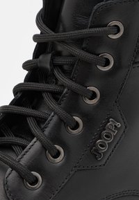 Bottine en cuir noir avec bout rond, œillets métalliques, lacets noirs et un détail de marque. Texture lisse et construction robuste.