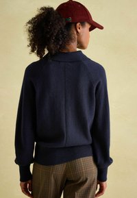 Marineblauer Strickpullover mit geripptem Saum, Raglanärmeln und weitem Kragen, kombiniert mit karierten Hosen. Der Hintergrund hat einen neutralen Farbton.