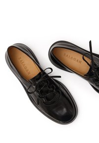 Scarpe in pelle verniciata nera con punta arrotondata, caratterizzate da un motivo decorativo in pizzo e una superficie liscia. All'interno, è visibile una soletta beige.