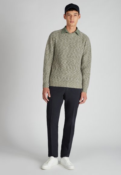 Un jeune homme portant un pull en tricot vert par-dessus une chemise à col verte, un pantalon noir, des baskets blanches et une casquette sombre sur un fond uni.