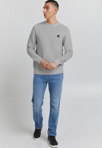 Felpa grigia a coste con collo rotondo e una piccola applicazione nera, abbinata a jeans azzurri chiari e scarpe nere, in piedi su uno sfondo semplice.