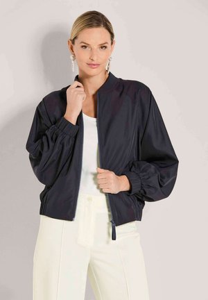 Frau trägt übergroße schwarze Bomberjacke über weißem Top und cremefarbenen Hosen, posiert vor einfarbig hellem Hintergrund.