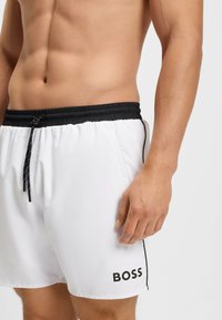 Férfi torso fehér úszóshortban, fekete derékpánttal, húzózsinórral és bal lábon "BOSS" logóval, egyszínű világos háttér előtt.