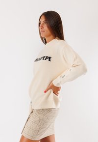 Maglione a maglia crema con scollatura rotonda e testo nero "TRITAPEPE." Presenta polsini e orlo a coste, abbinato a una gonna a quadri beige.