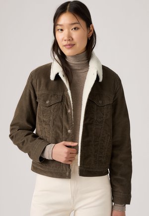 Junge Frau trägt eine braune Cordjacke mit weißem Fleecekragen und taupefarbenem Rollkragenpullover, stehend vor einem schlichten hellen Hintergrund.