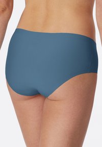 Donna che indossa biancheria intima blu senza cuciture a vita alta, vista da dietro, su sfondo bianco uniforme.