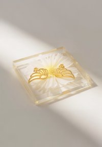 Orecchini a forma di farfalla in oro posati su una margherita bianca all'interno di una vetrina quadrata di resina trasparente. Superficie texturizzata e bordi lisci messi in evidenza.