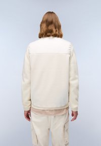 Weiße Fleecejacke mit einem glatten, hellbeigen Stoffübergang, gerippten Bündchen und einer lockeren Passform. Getragen über beigen Hosen.