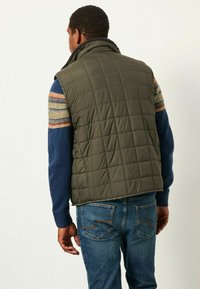 Olivgrüne gesteppte Weste mit hohem Kragen und strukturiertem Futter, getragen über einem blauen Musterpullover, kombiniert mit blauen Jeans.