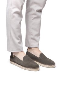 Hallid hall suède slip-on kingad elastse avausega, millel on kootud jute tald ja tikand ääre ümber. Kannab heledat värvi pükse.