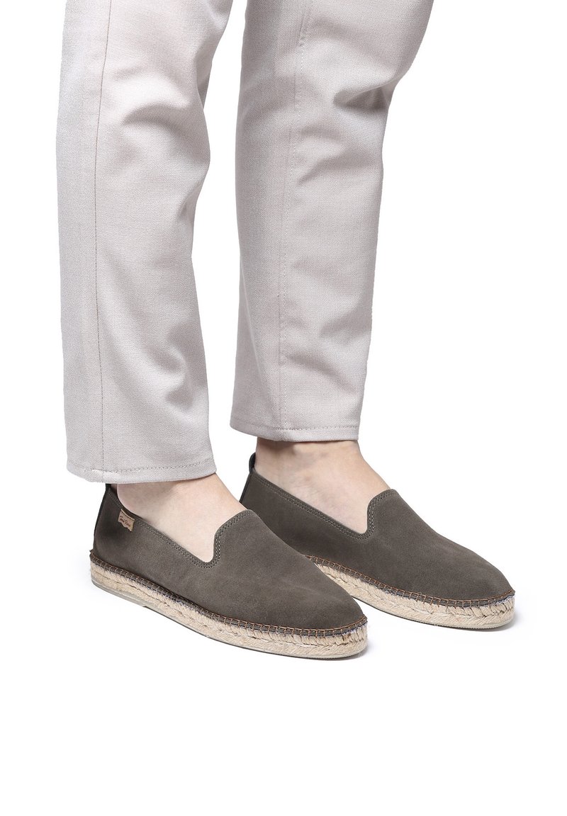 Hallid hall suède slip-on kingad elastse avausega, millel on kootud jute tald ja tikand ääre ümber. Kannab heledat värvi pükse.