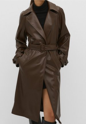 Mujer con un abrigo trench de cuero sintético marrón oscuro con cinturón y solapas anchas, sobre un conjunto negro y botas negras. Manos en los bolsillos del abrigo.