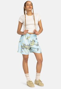 Shorts à motifs floraux bleu clair avec une taille élastique, associés à un crop top beige ajusté, des chaussettes blanches et des chaussures beiges.