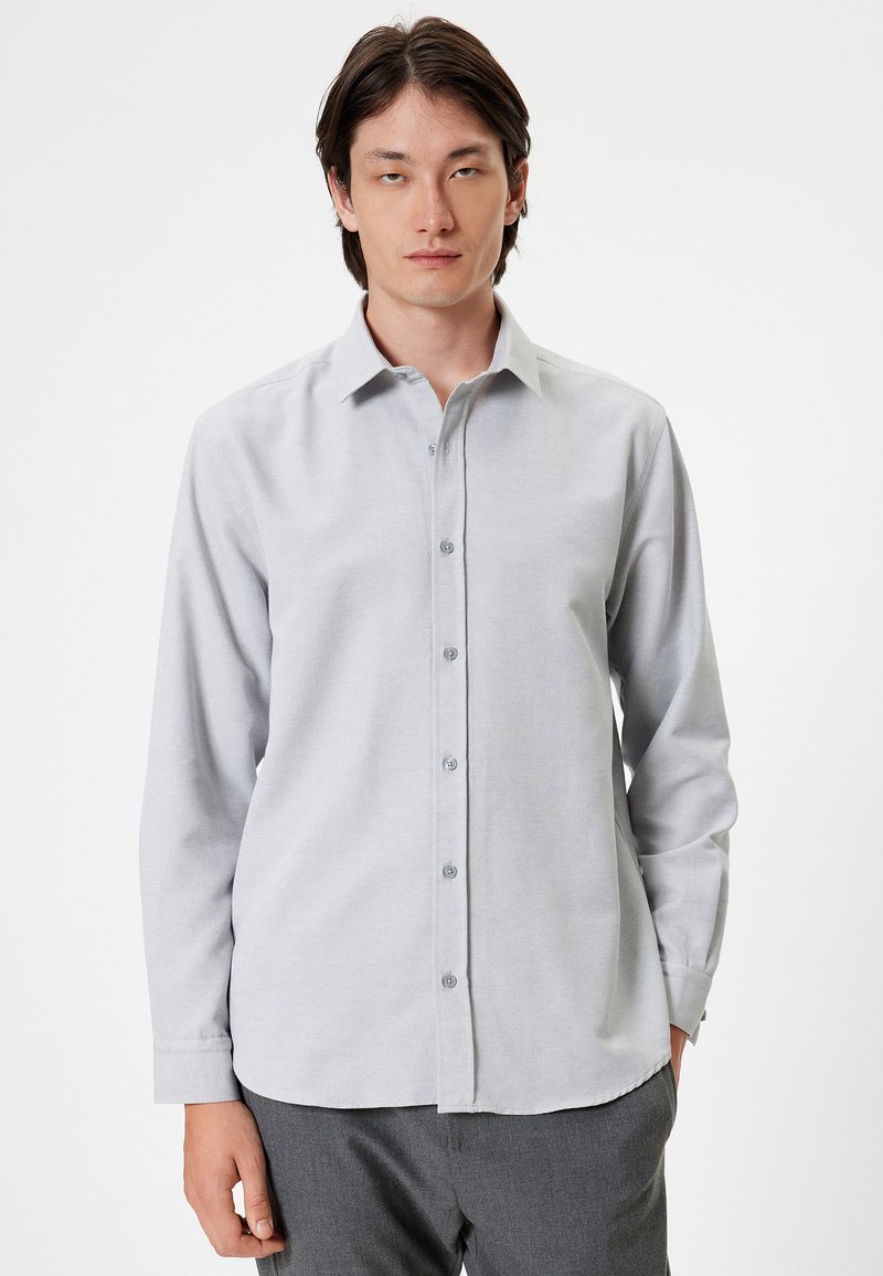 Lys grå skjorte med lange ærmer, krave og lige kant. Har matchende knapper og en glat tekstur.