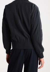 Chaqueta bomber negra, ligera, con dobladillo y puños elásticos, que presenta una textura suave y un diseño minimalista sin cierres visibles.