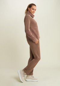 Donna in piedi di lato con le mani nelle tasche, indossa un top marrone a maniche lunghe, pantaloni abbinati e sneakers con piattaforma foderate in pelliccia beige.