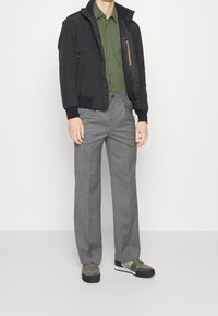 Černá bomber bunda se zipovými detaily, zelená košile s límcem, šedé kalhoty a olivové tenisky s bílou podrážkou a námořnickými akcenty.