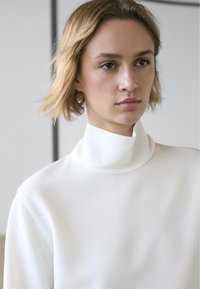 Bíl�ý top s vysokým límcem a hladkou texturou, s dlouhými rukávy a minimalistickým designem. Látka působí lehce a jemně.