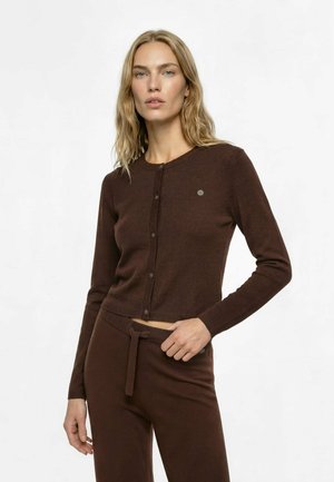 Frau mit blonden Haaren, die einen braunen, geknöpften Cardigan und passende Hose mit Kordelzug trägt, steht vor einem schlichten hellen Hintergrund.