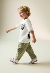 Weißes Baumwoll-T-Shirt mit Grafikdruck, kombiniert mit grünen karierten Hosen und weißen Sneakern mit roten Akzenten. Kind läuft auf hellem Hintergrund.