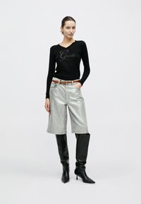 Top negro de manga larga con el logo de "Guess", pantalones cortos plateados hasta la rodilla, cinturón marrón y botas negras puntiagudas hasta la rodilla. Fondo neutro.