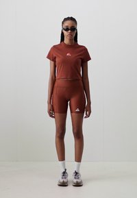 Brūns īsais krekls ar logo, atbilstoši augstie šorti un baltās sporta zeķes. Modeli nēsā melni saulesbrilles un sporta apavus.