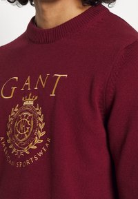 Pull bordeaux avec un col rond, arborant un logo et un texte brodés en or sur la poitrine. Le tissu a une texture lisse et des poignets côtelés.