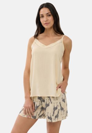 Kvinde iført en ærmeløs beige top og beige shorts med sorte giraftryk, stående med den ene hånd i lommen mod en ensfarvet baggrund.