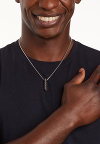 Zilveren ketting met een rechthoekige zwarte hanger waarop een rij kleine, ronde versieringen is te zien; gedragen op een donker shirt.