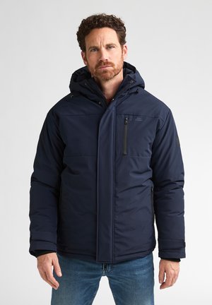 Man wearing a navy blue hooded winter jacket with zipper pockets and blue jeans standing against a plain background.