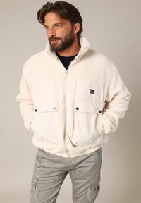 Veste en polaire couleur crème avec un devant en tissu lisse, fermeture éclair, deux grandes poches à rabat et un col haut.